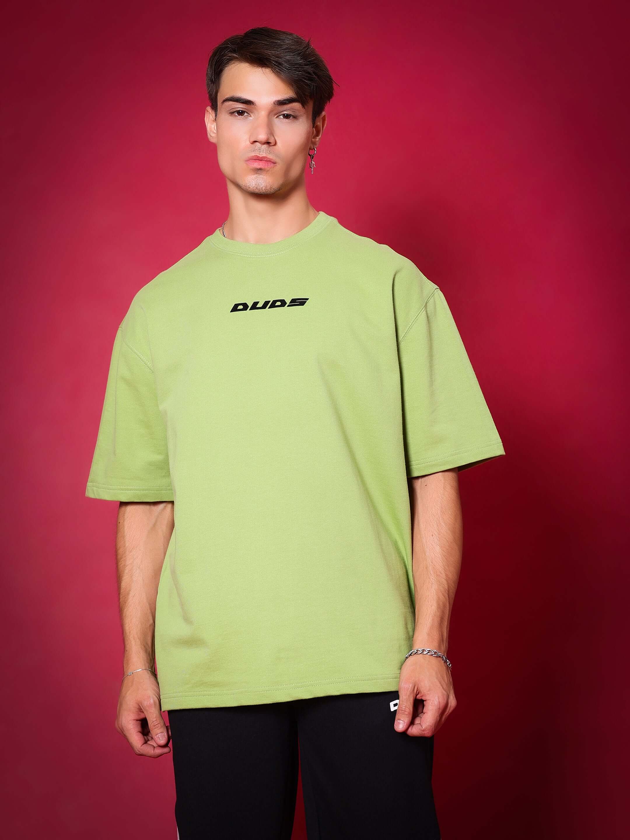 Model wearing Scorpio oversized T-shirt in pista green, premium cotton French terry fabric, photographed against a red background.