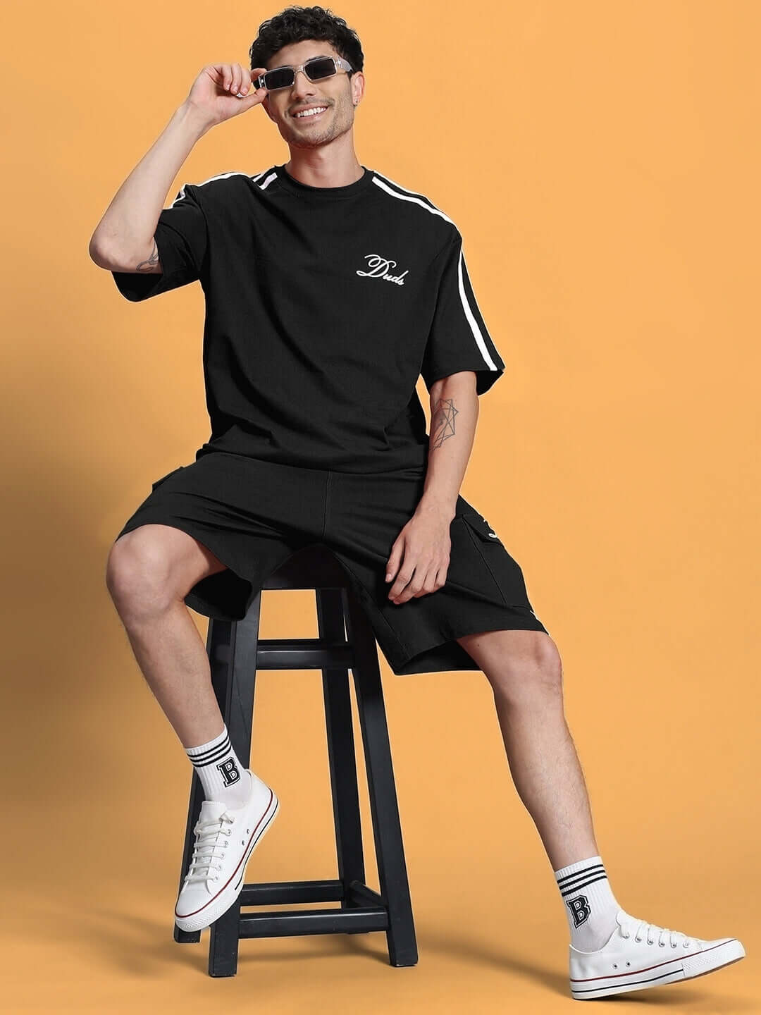 Model wearing SCOTIA SUMMER CO-ORD SET BLACK, featuring cotton terry fabric, sitting on a stool with casual sneakers.