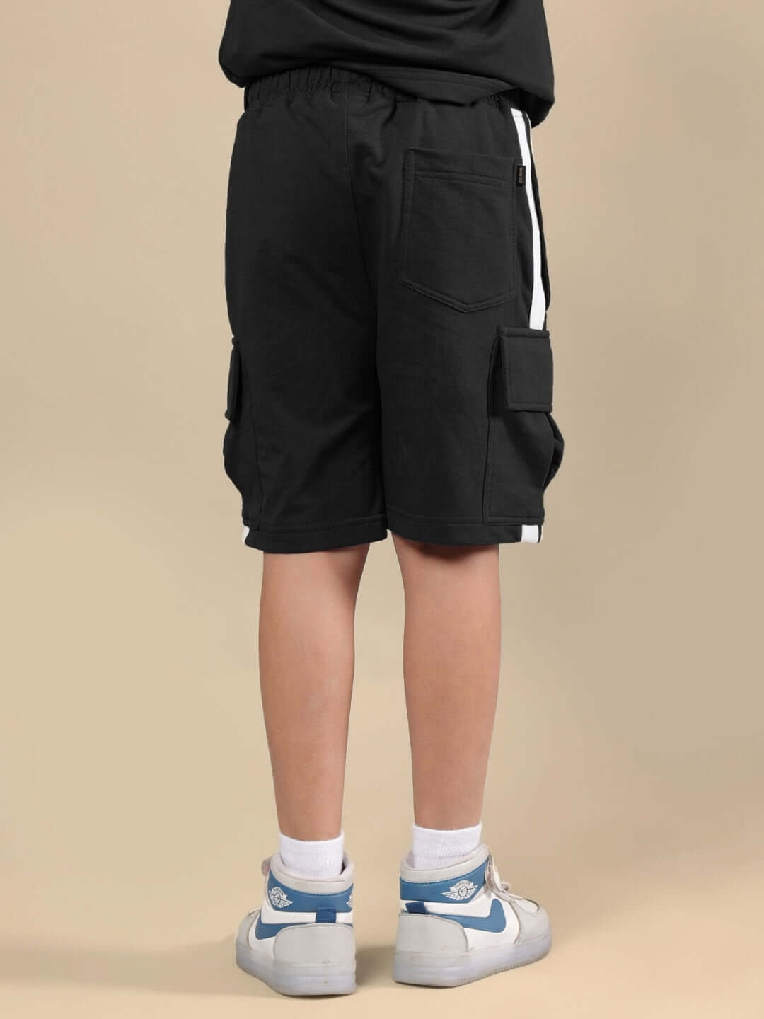 Child wearing SCOTIA SHORTS FOR BOYS & GIRLS BLACK with white sneakers; back view showing pocket and design details.