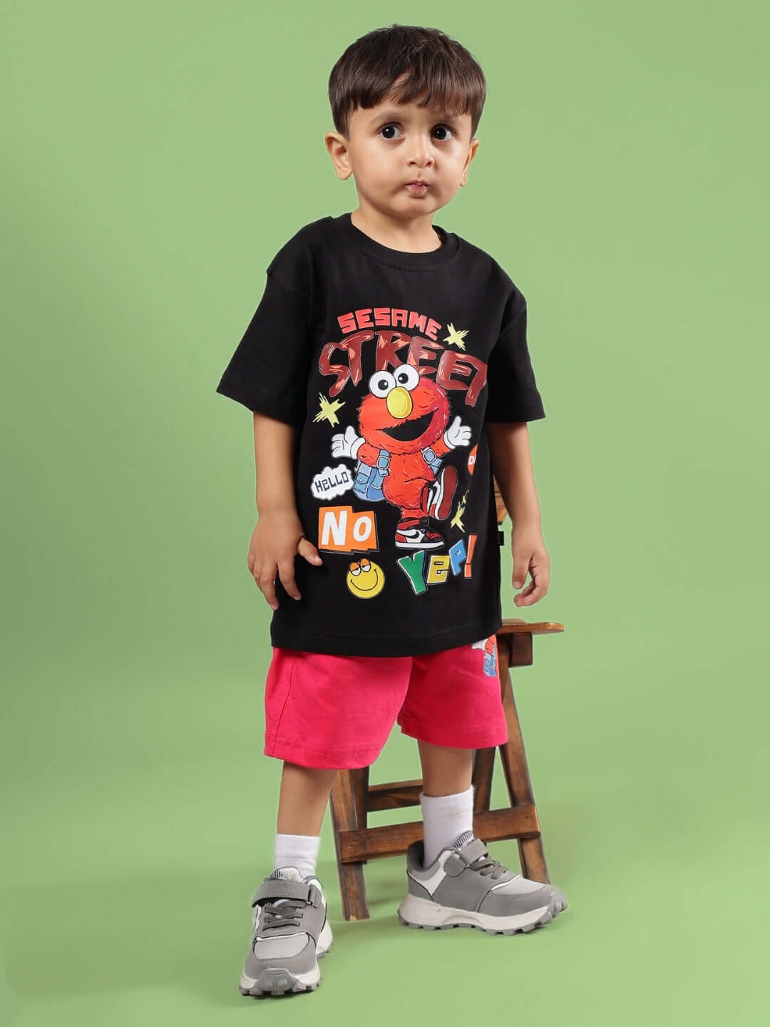 Child wearing black Sesame Street shirt and pink shorts with sneakers, standing in front of a green background.