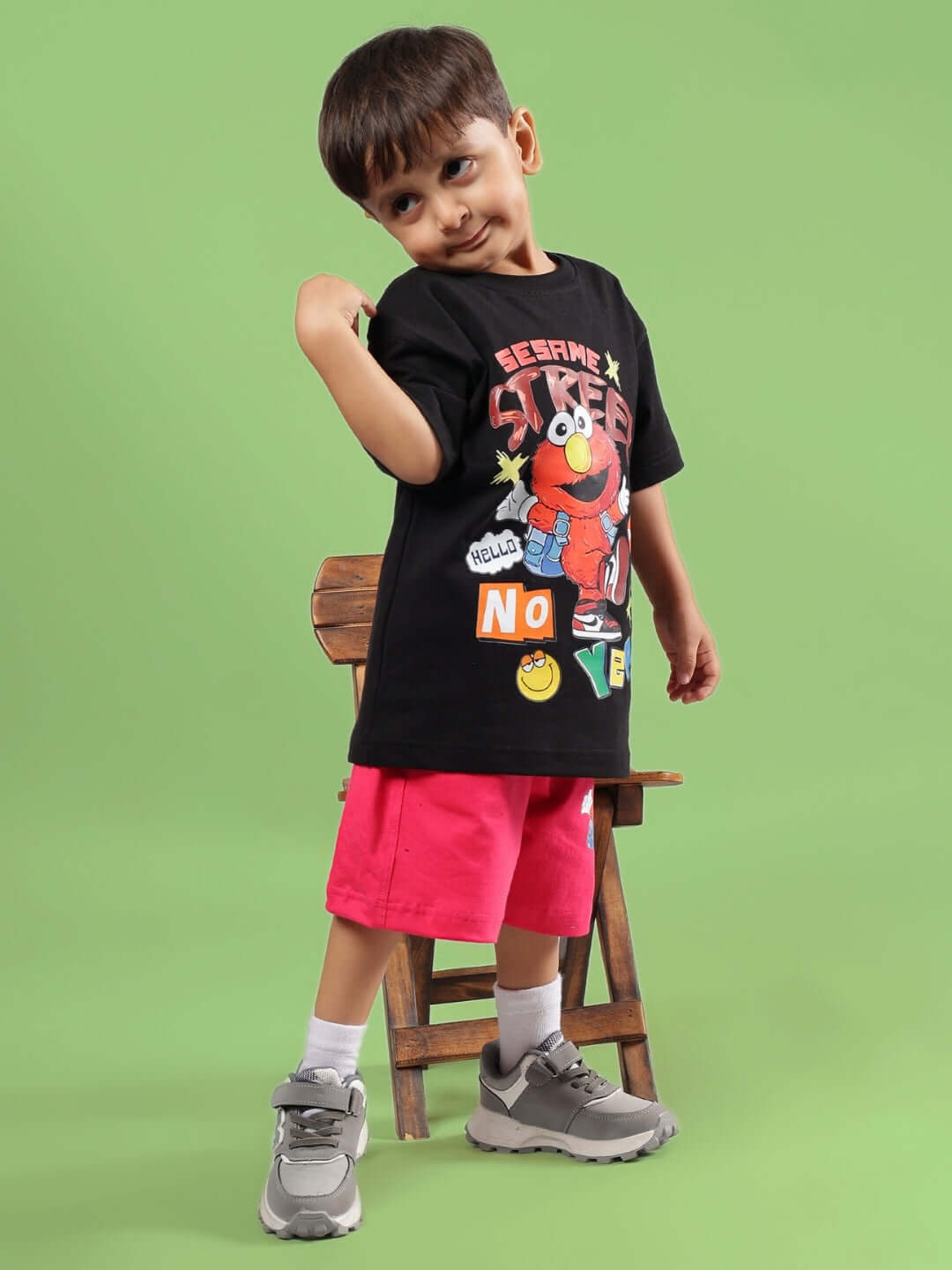 Child wearing a black Sesame Street-themed T-shirt and pink shorts from DUDS, posed against a green background.