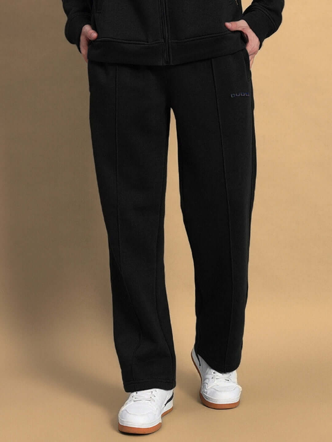 Person wearing SHAGGY ZIPPER FLEECE CO-ORD SET BLACK showing detail of pants with white sneakers on beige background.