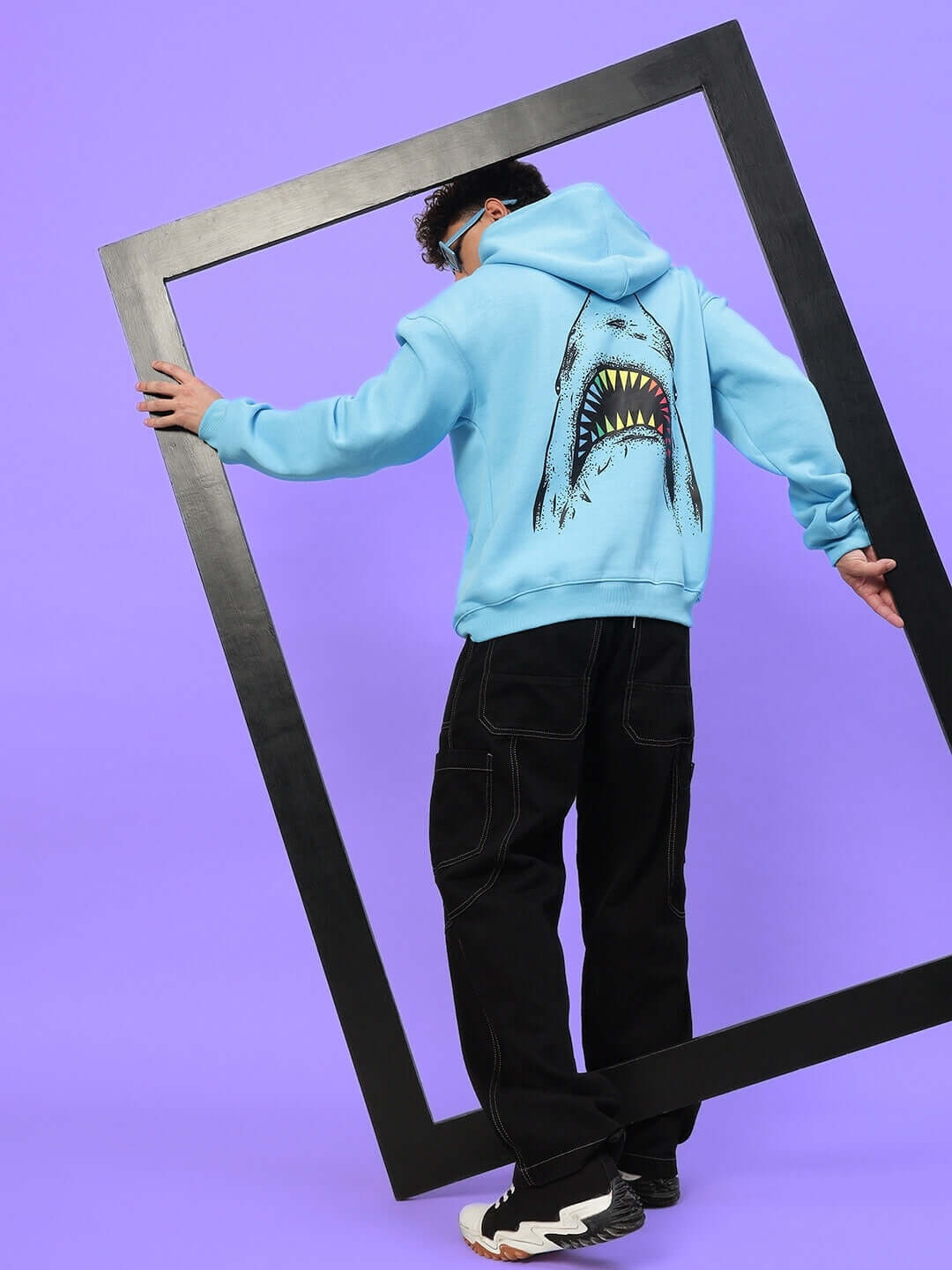 Model in SHARKWAVE OVERSIZED FLEECE HOODIE LIGHT BLUE with shark graphic, holding black frame on purple background.