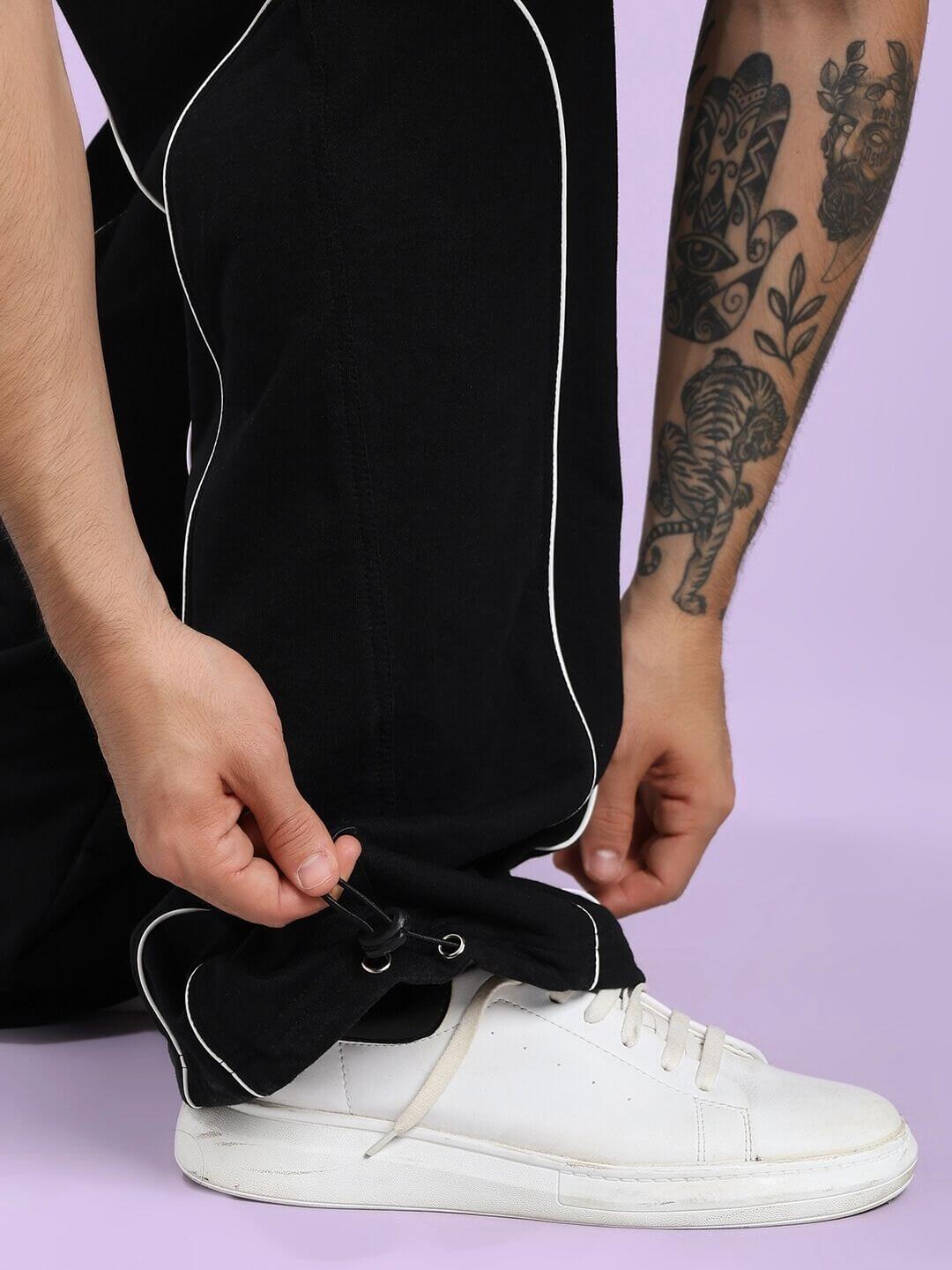 Person adjusting SIGNATURE PIPING JOGGERS BLACK with white sneakers, showcasing stylish contrast piping and comfortable fit.