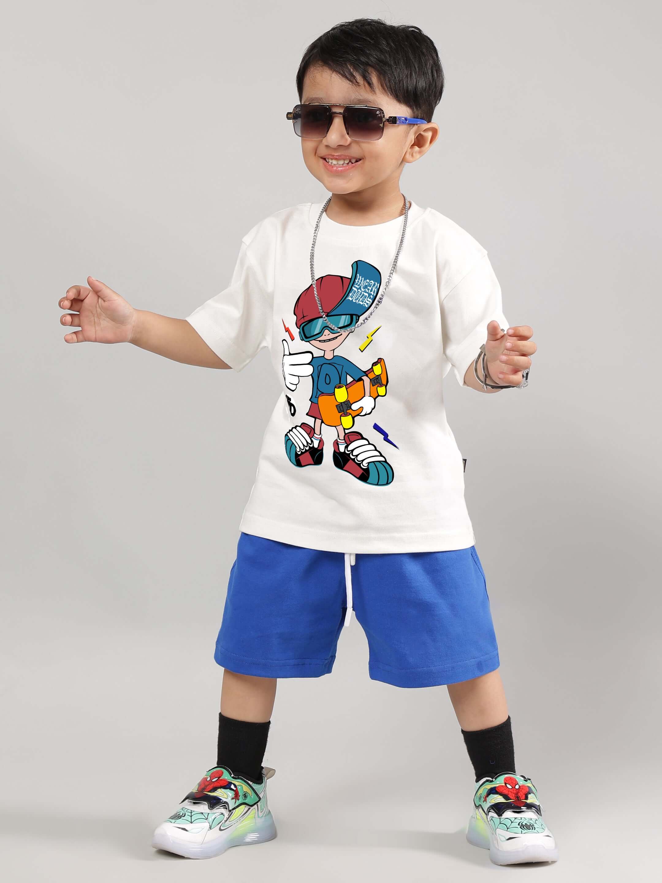 Boy wearing SKATE RIDER T-shirt in off white, featuring a colorful skateboard graphic, paired with blue shorts and playful shoes.