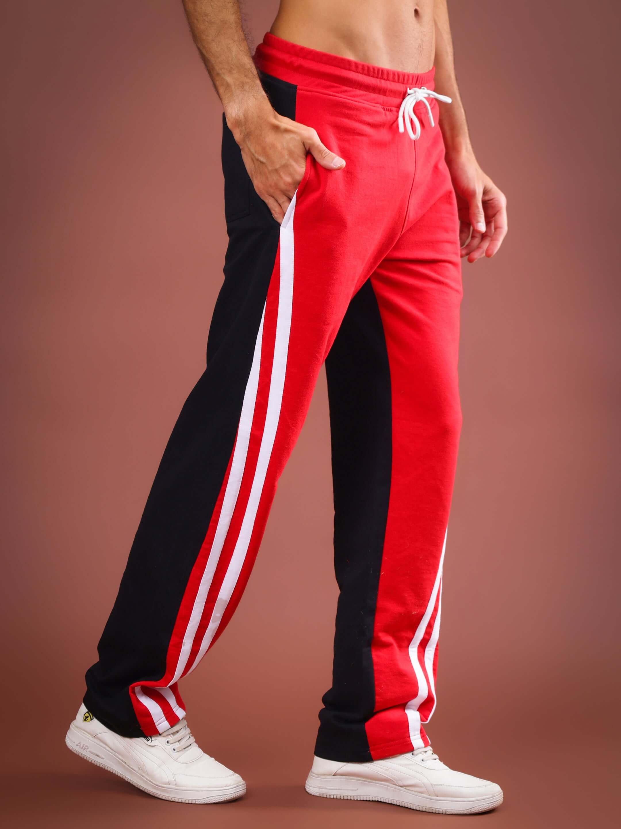 Man wearing red and black skater baggy joggers with white sneakers, featuring side stripes, modeled against a brown background.