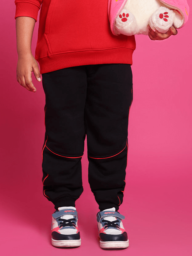 Child wearing black relaxed fit joggers with red accents, holding a plush toy, against a pink background.