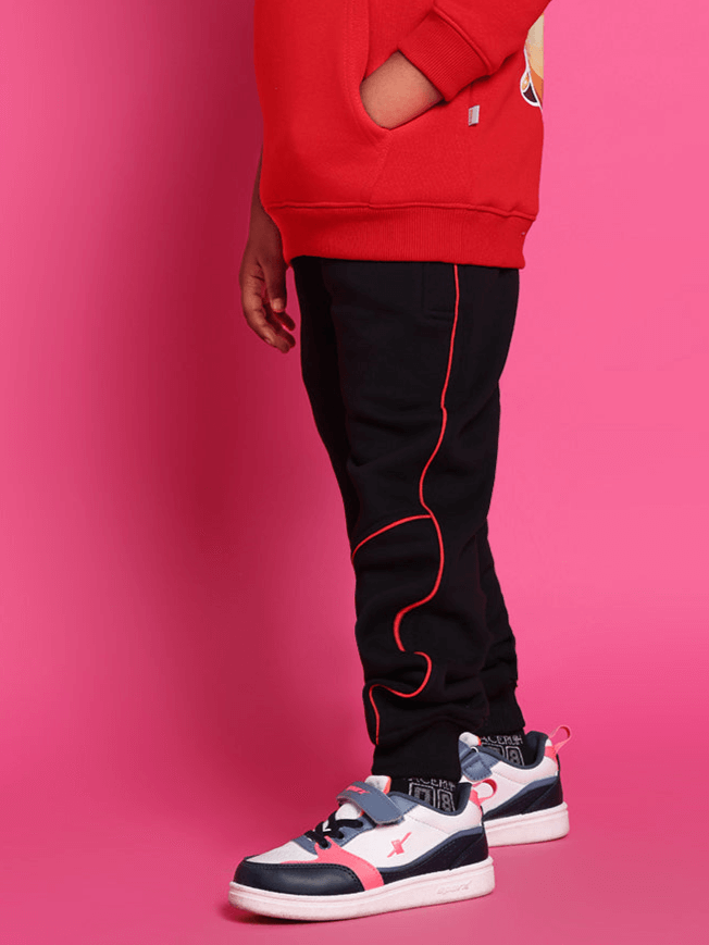 Child wearing black relaxed fit joggers with red piping, paired with sneakers against a pink background.