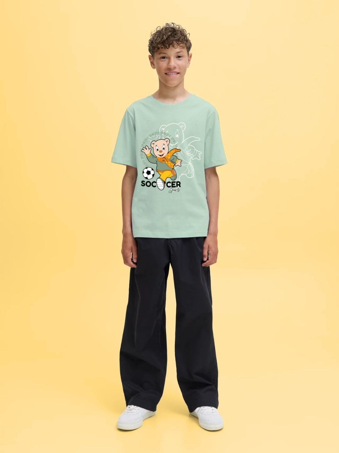 Boy wearing SOCCER PRINTED T-SHIRT FOR BOYS AND GIRLS LIGHT GREEN with soccer graphic on light green background.