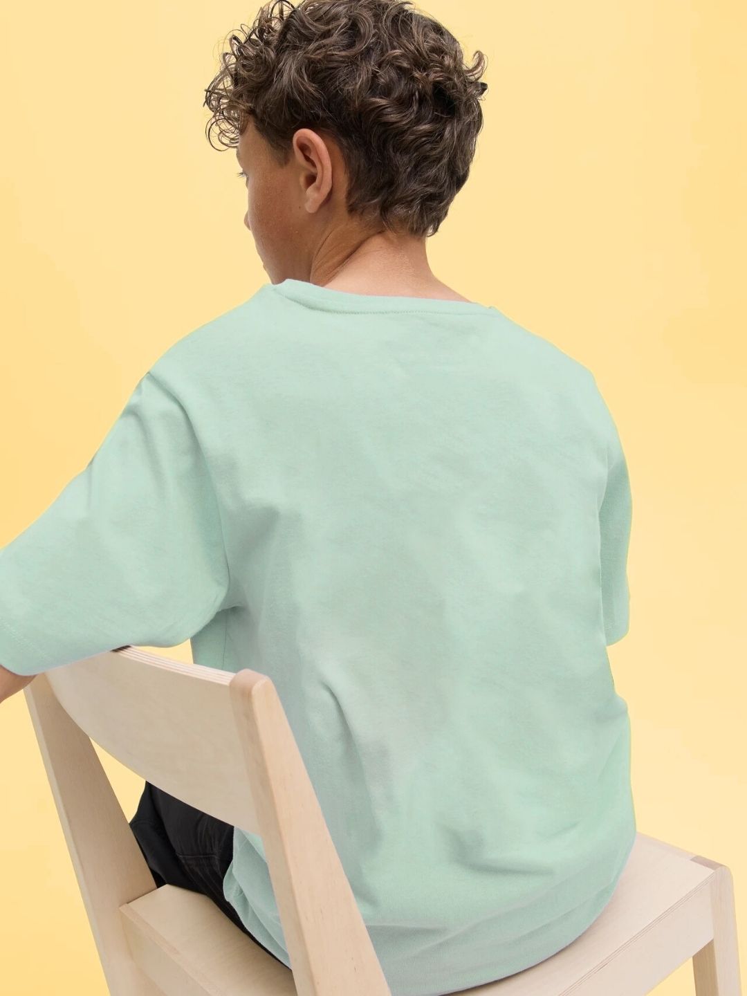 Child wearing SOCCER PRINTED T-SHIRT FOR BOYS AND GIRLS LIGHT GREEN sitting on a chair, showcasing the back design against a yellow background.