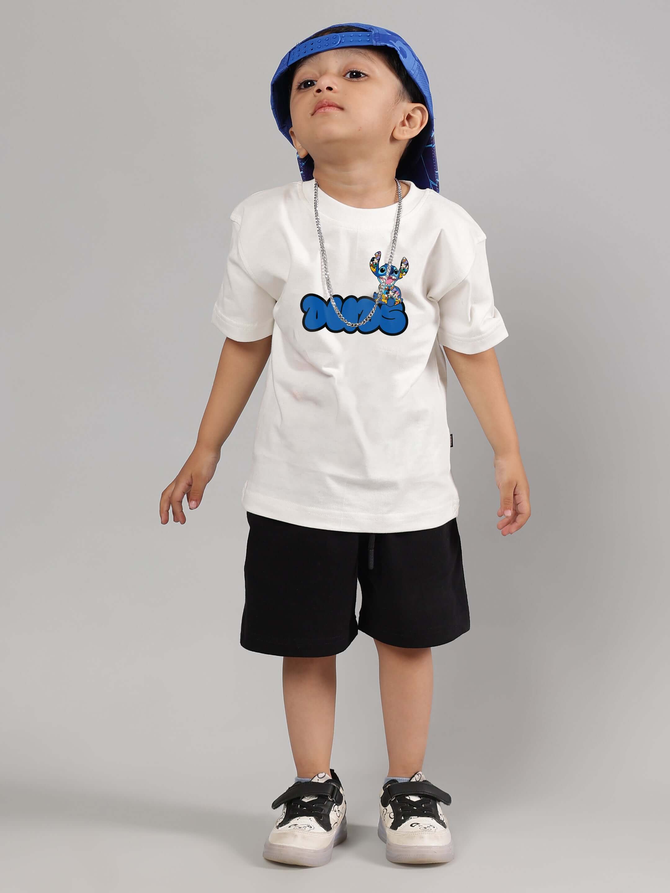 Child wearing SORE SUMMER CO-ORD in off white and black, featuring graphic tee and shorts, styled with a blue hat and sneakers.