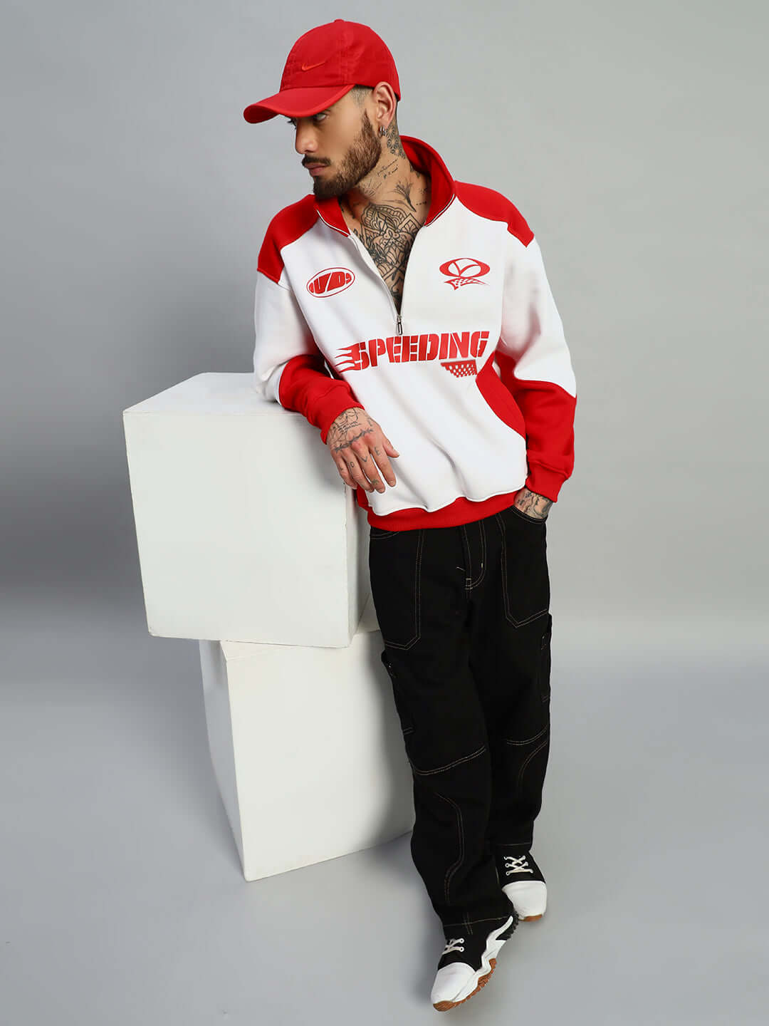 Man wearing a red and white SPEEDING colorblock jacket, leaning on white geometric blocks, paired with a red cap and black pants.