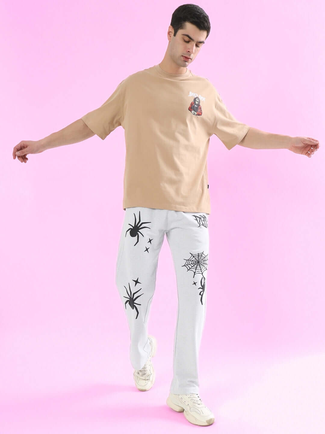 Man wearing light grey joggers with spider web design, styled with a beige t-shirt, walking on a pink background.