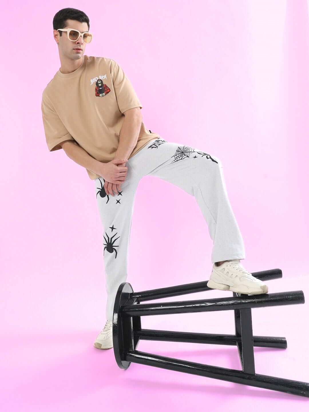 Model wearing light grey joggers with spider web design, standing on a black stool, against a pink background. Perfect casual wear.