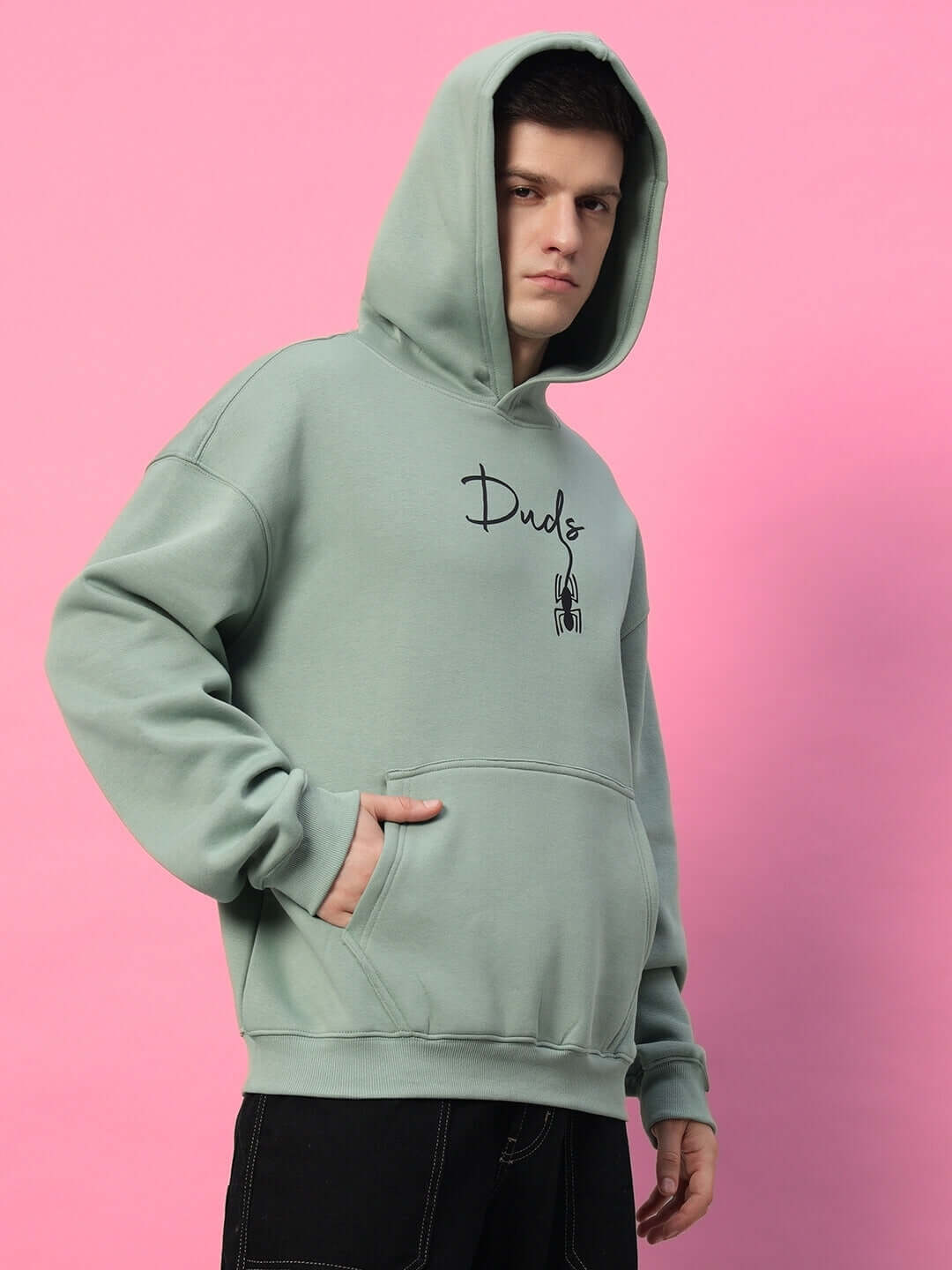 Adult wearing a SPIDERMAN OVERSIZED FLEECE HOODIE JADE GREEN against a pink background, featuring a hood and hand pocket.