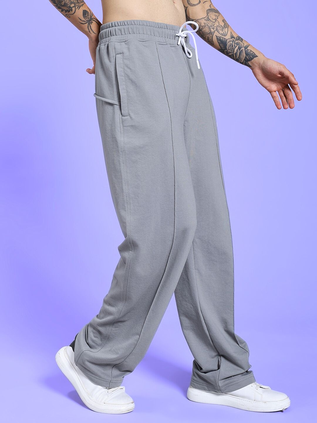 Model wearing SPIRAL RELAXED-FIT JOGGERS GREY with tattoos and white sneakers against purple background