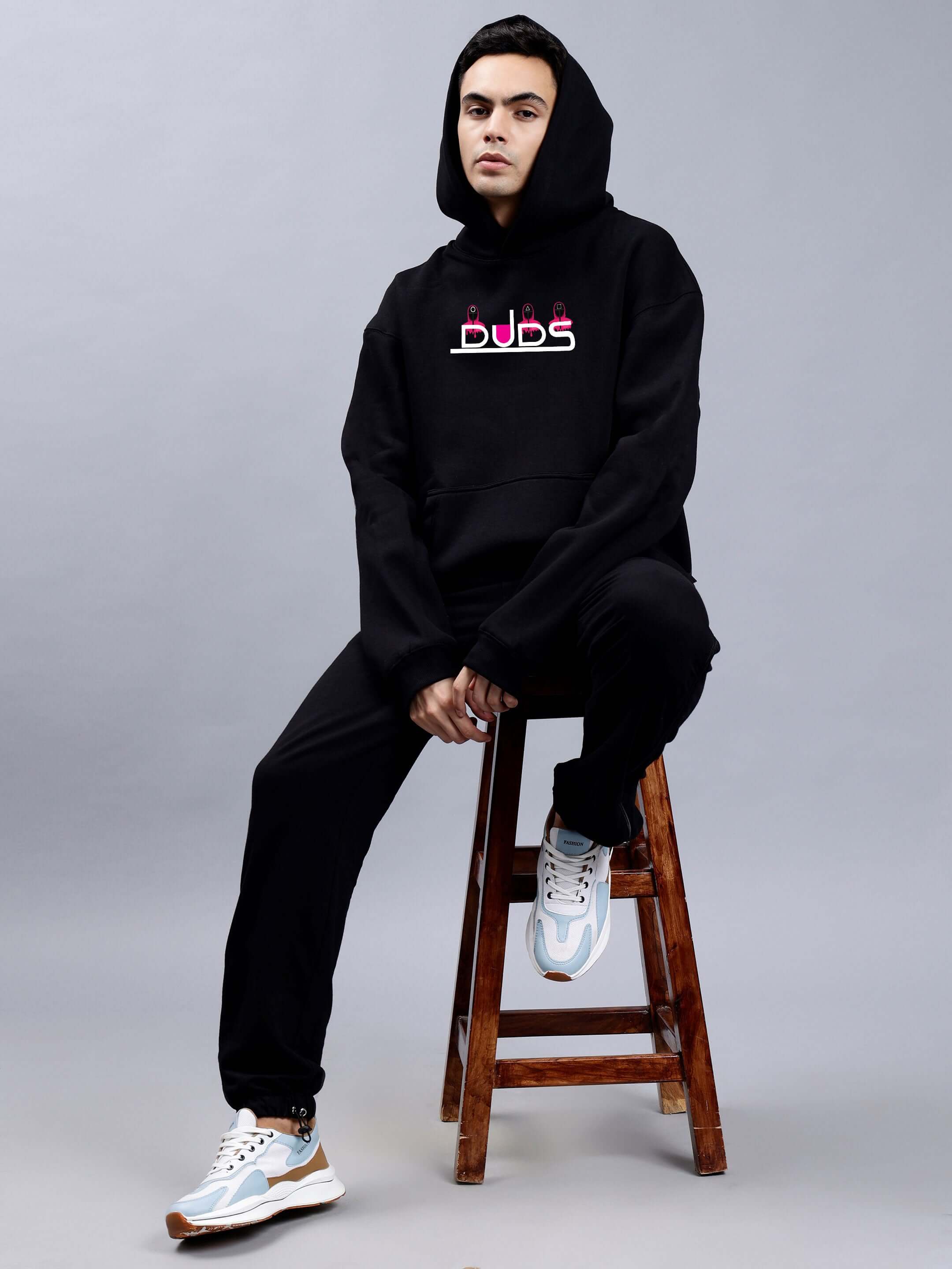 Model wearing Squid Game Oversized Hoodie Men Black, sitting on stool, showcasing relaxed fit and streetwear style.