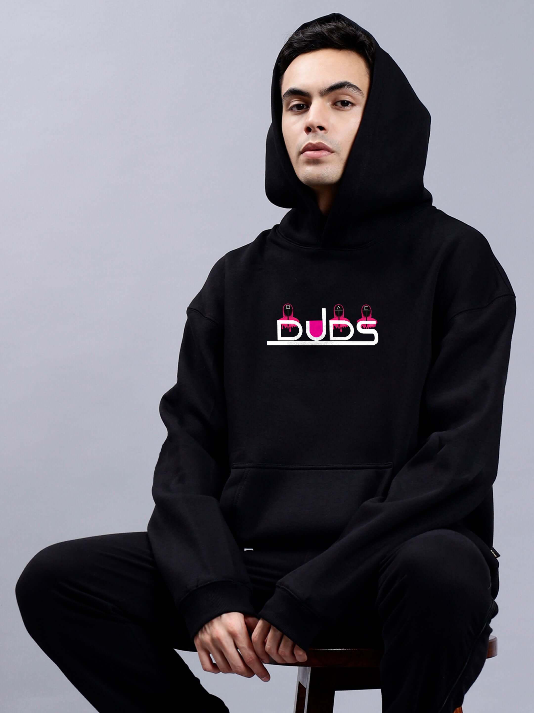 Man wearing SQUID GAME OVERSIZED HOODIE MEN BLACK with logo, seated in a relaxed pose.