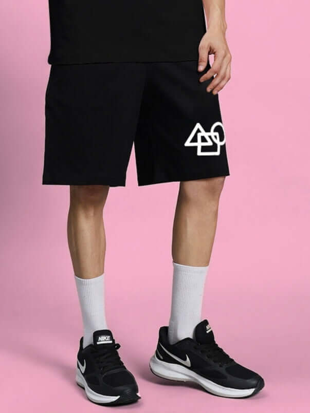 Man wearing SQUID GAME SUMMER SHORTS BLACK with geometric design, standing against a pink background.