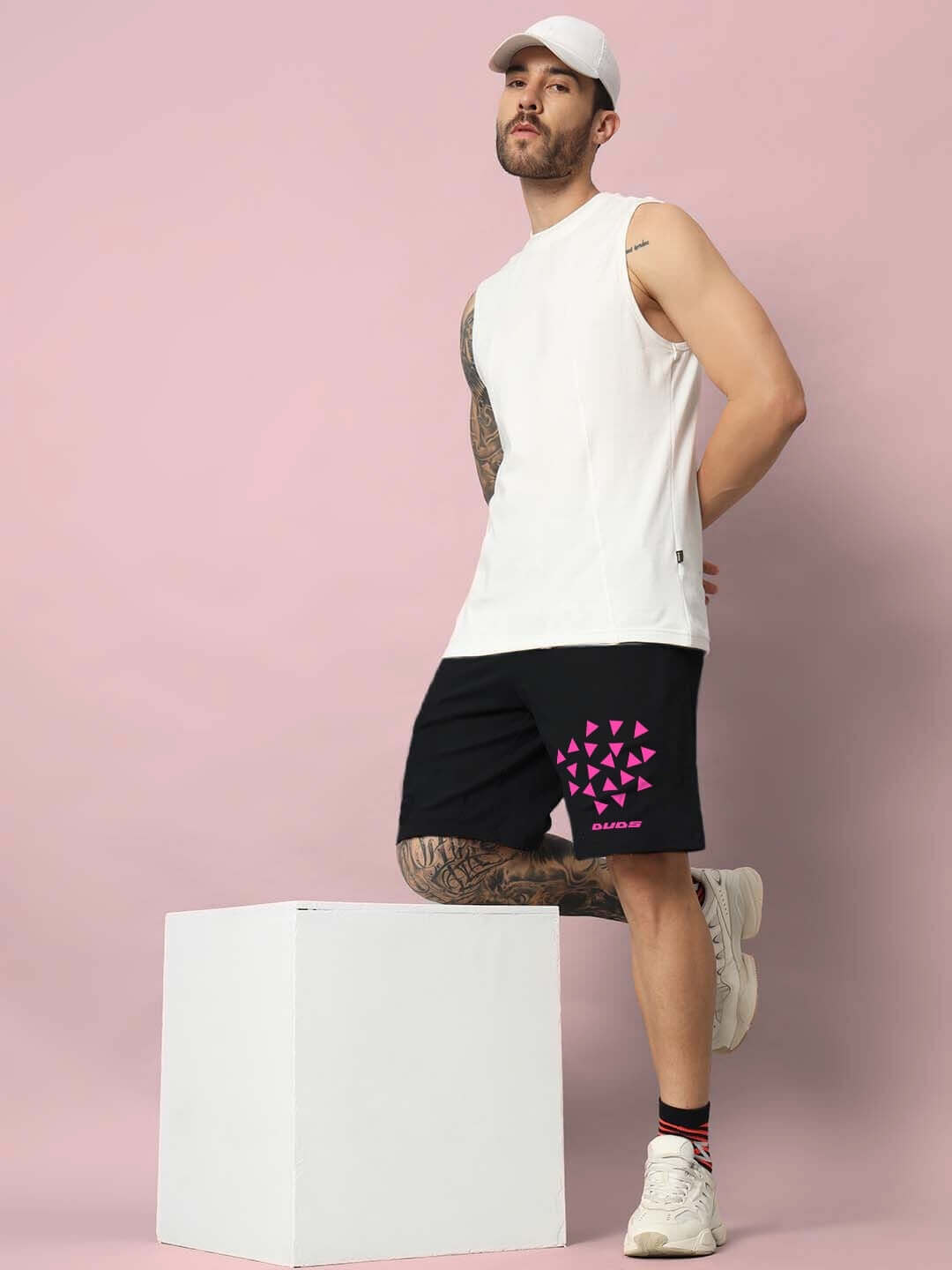 Man wearing STAR DUST Regular Fit Shorts in black with pink geometric pattern, paired with a white tank top and cap, posing against a pink background.