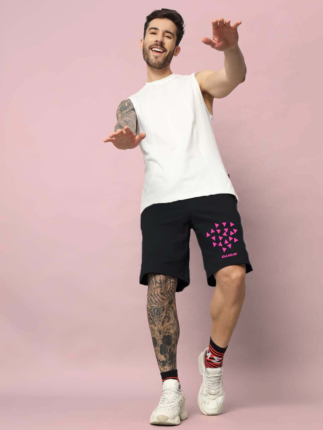 Man wearing STAR DUST regular fit black shorts with pink design, white sleeveless shirt, showcasing casual style against a pink background.