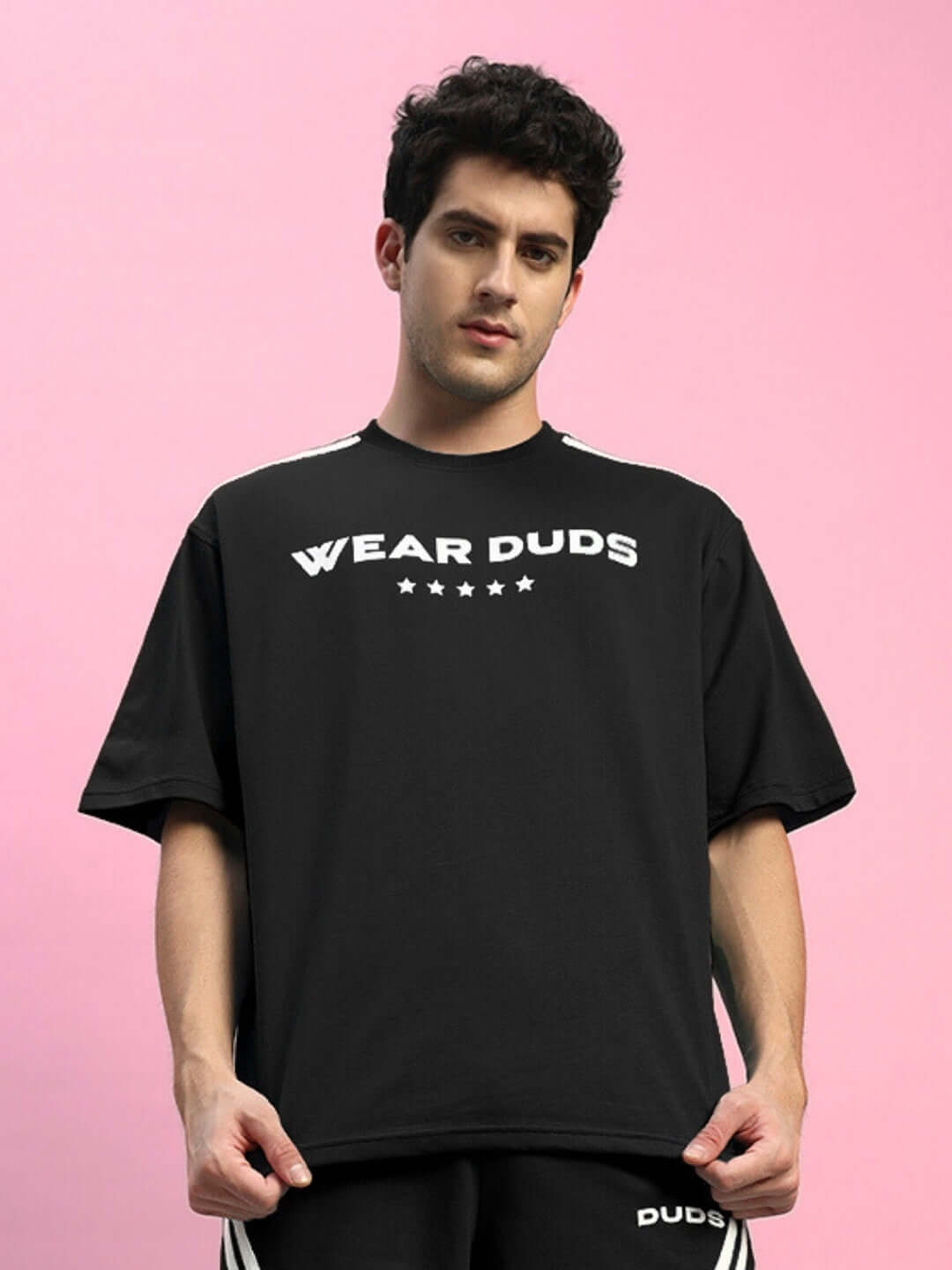 Model wearing STAR JUMPER OVER-SIZED T-SHIRT BLACK, showcasing casual streetwear style against a pink background.