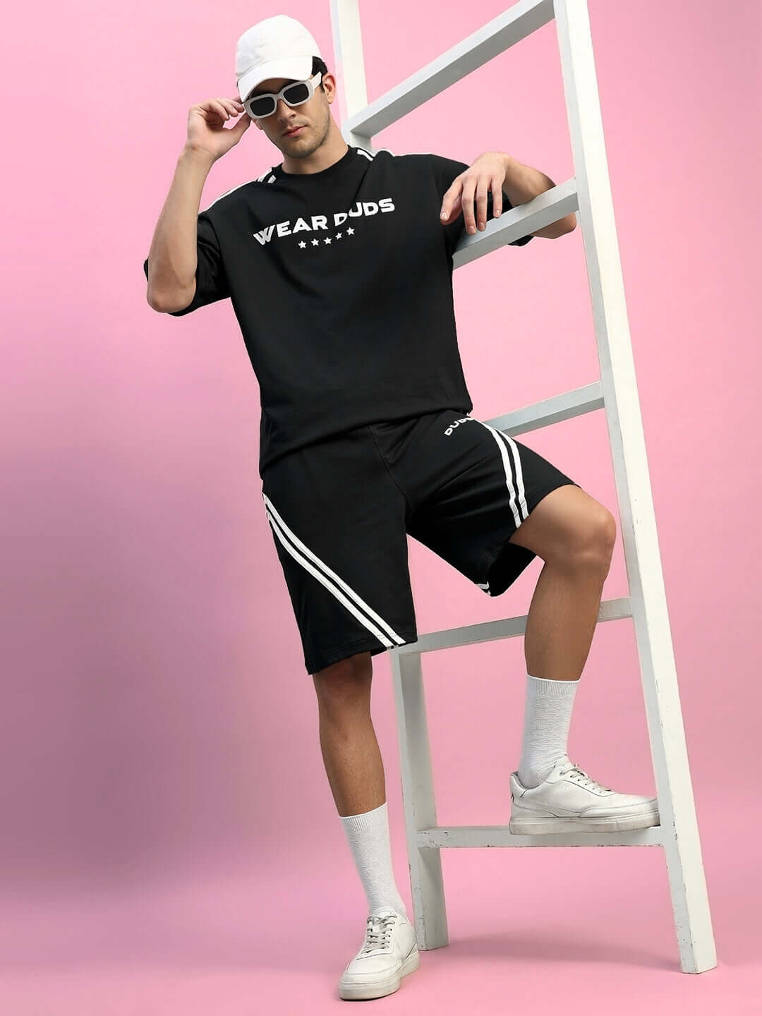 Model wearing black STAR JUMPER regular fit shorts with custom fit option, standing on a ladder against a pink backdrop.