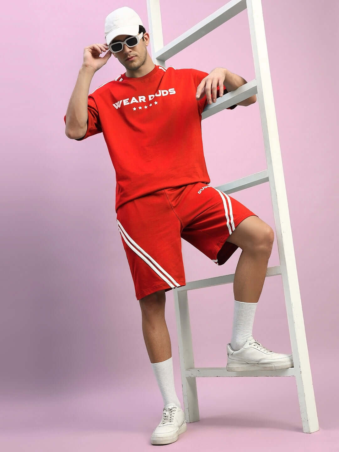 Model wearing red STAR JUMPER Regular Fit Shorts, with white stripe detail, styled with matching shirt and sneakers against a pink background.