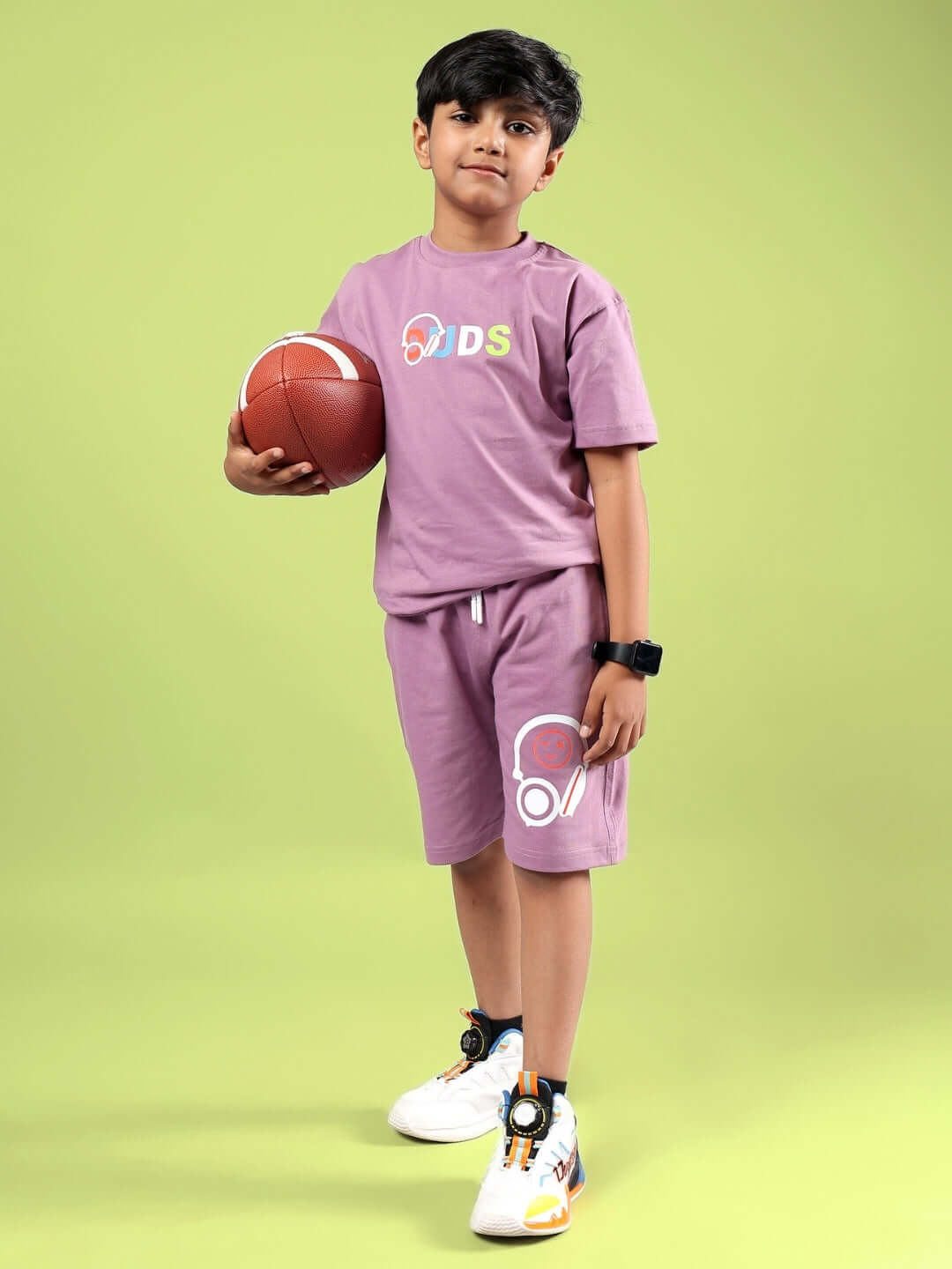 Young boy wearing STOIC SHORTS FOR BOYS & GIRLS PULM PURPLE standing with a basketball on green background.