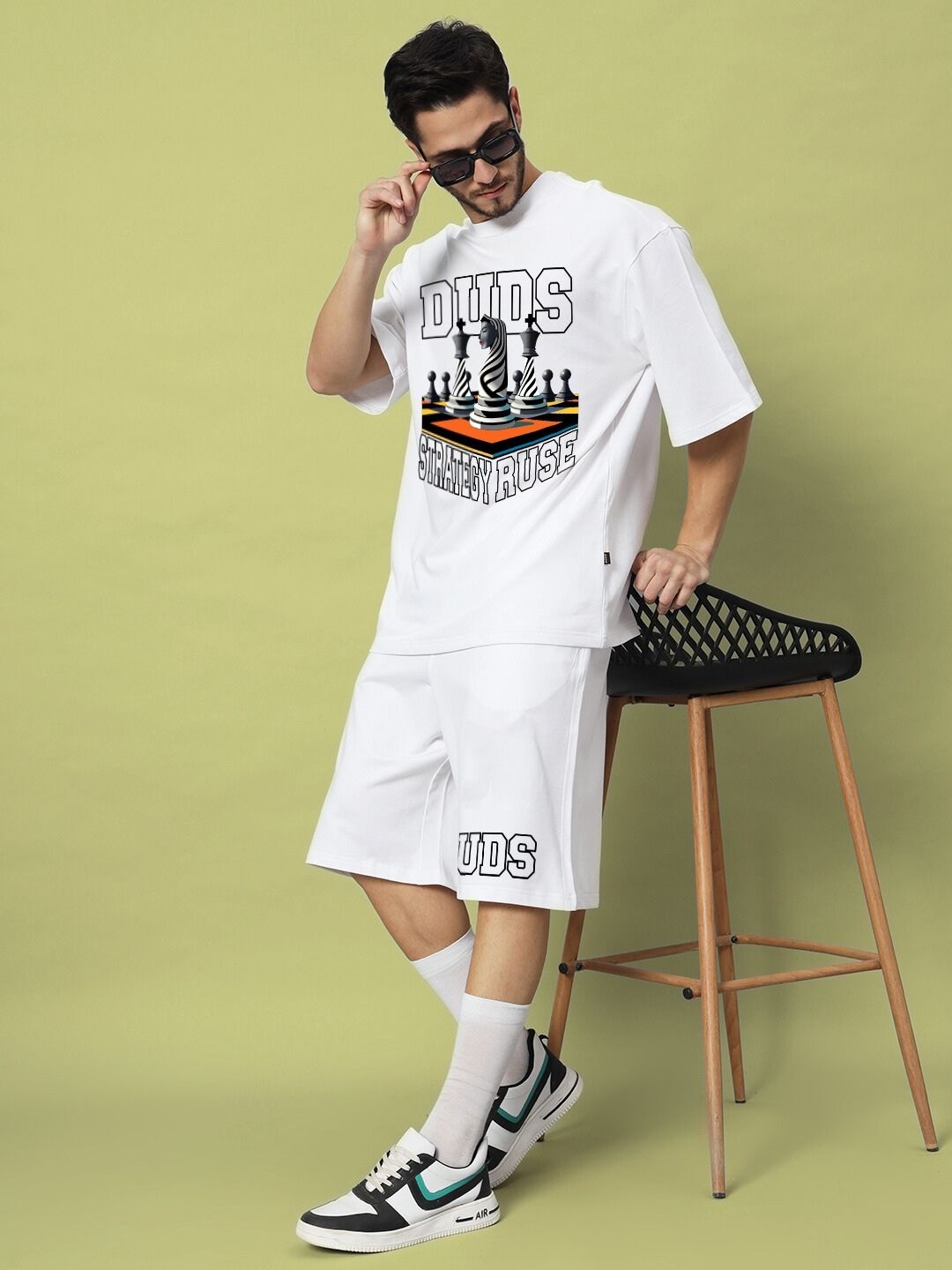 Man modeling STRATEGY RUSE CO-ORD SET WHITE with graphic design, featuring shorts and t-shirt, leaning on a stool against a light green background.