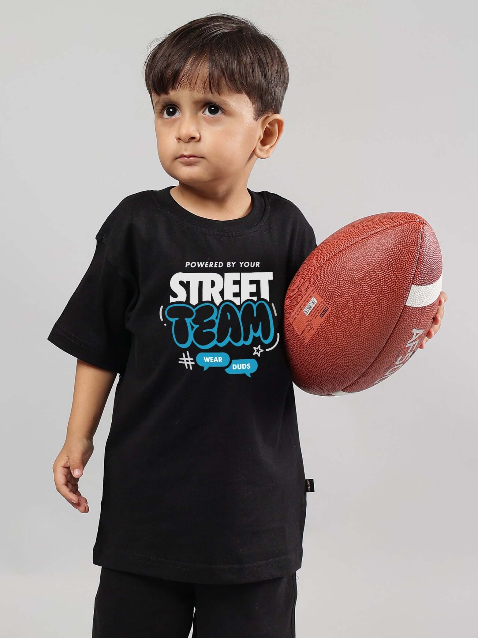 Young child wearing a black STREET TEAM T-shirt holding a football, showcasing DUDS apparel for boys and girls.