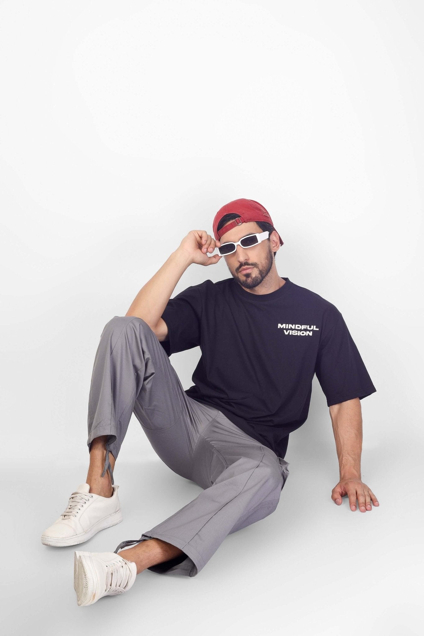 Man wearing Moon Stone Cargo Pants with 6 Cargo Pocket, black mindful vision t-shirt, red cap, and white sneakers sitting casually.