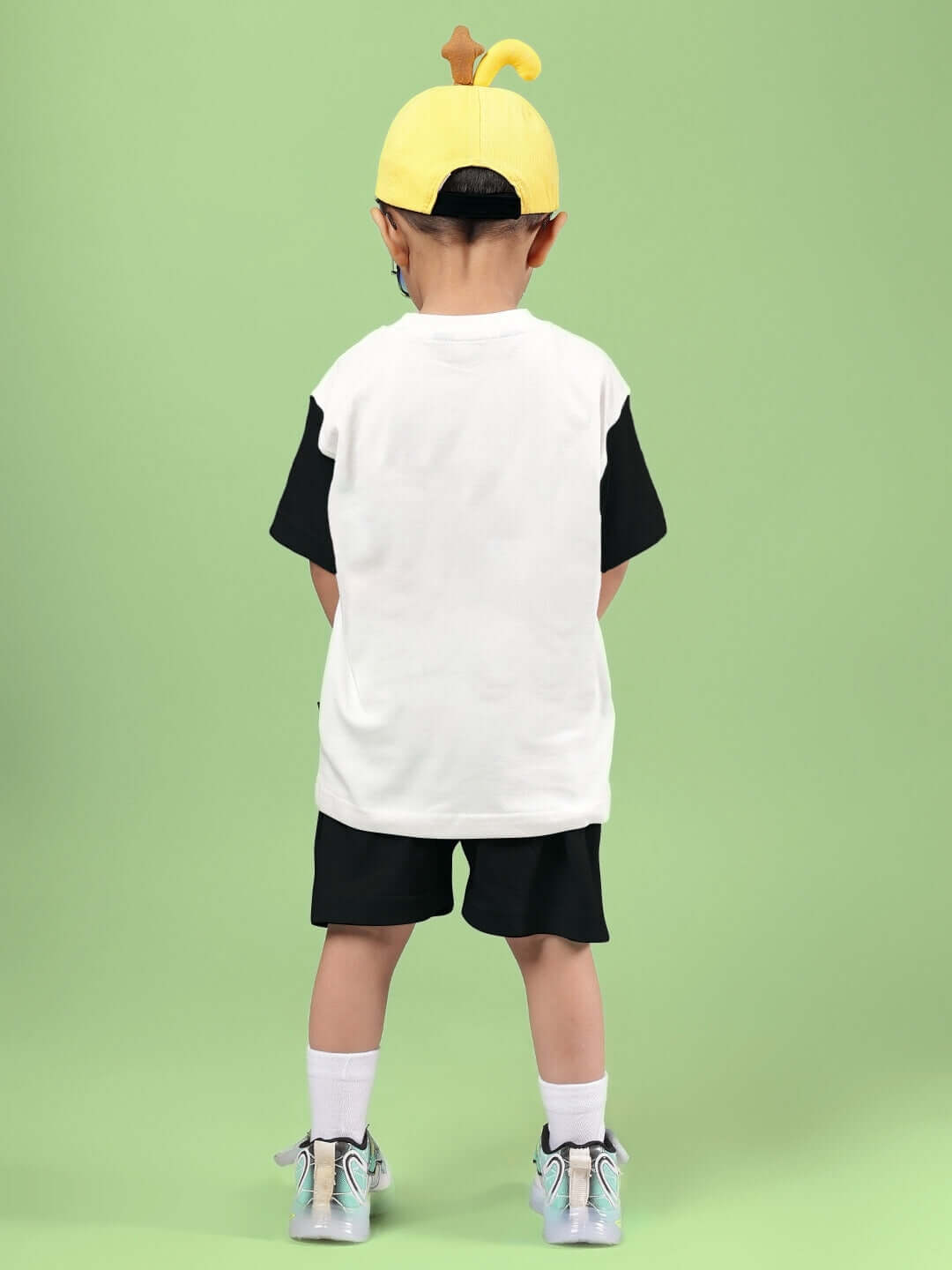 Child wearing off-white and black stylish robot co-ord set, back view, with a yellow cap on a green background.