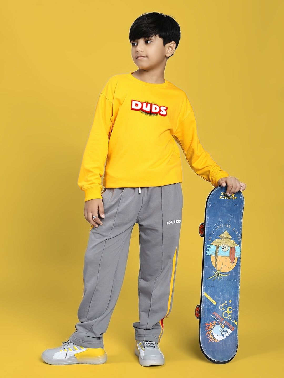 Child wearing SUNSHINE STRIPE JOGGER FOR BOYS AND GIRLS RED GREY holding a skateboard, standing against a yellow background.