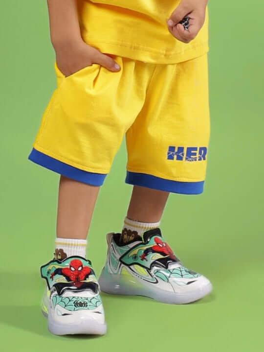 Child wearing SUPER HERO SHORTS FOR BOYS & GIRLS YELLOW with colorful sneakers on green background.