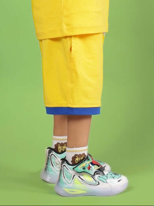 Child wearing SUPER HERO SHORTS FOR BOYS & GIRLS YELLOW with colorful sneakers, showcasing vibrant style on a green background.