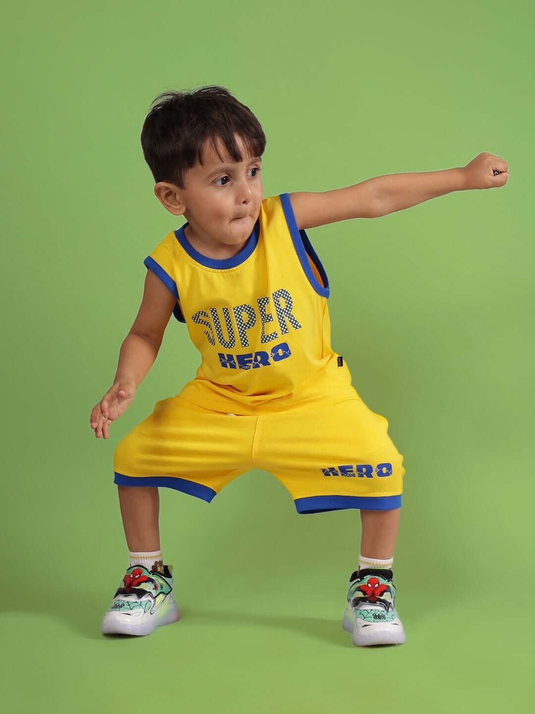 Young child wearing SUPER HERO SHORTS FOR BOYS & GIRLS YELLOW outfit striking a playful pose against a green background.