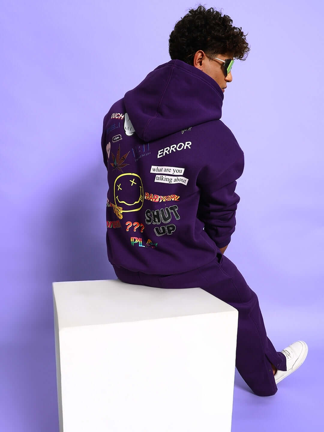 Person wearing SUPREME COTTON FLEECE CO-ORD PURPLE hoodie and joggers, featuring graphic prints and oversized fit, seated on a cube.