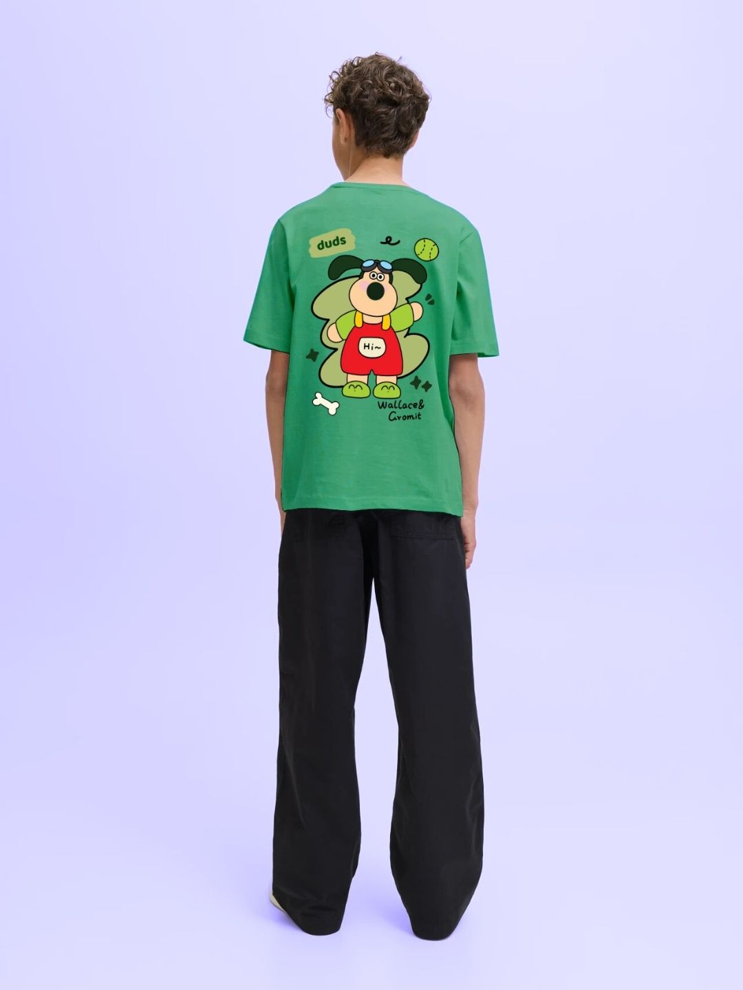 Person wearing TADDY PUPPY T-SHIRT FOR BOYS AND GIRLS GREEN with cartoon dog design, standing against light purple background.