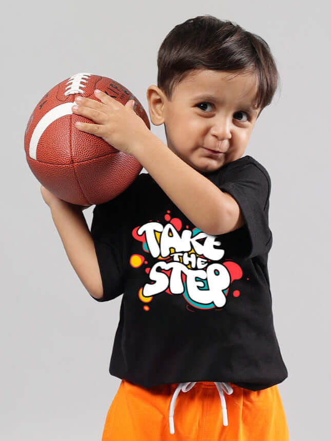 Young child holding a football, wearing a black "TAKE THE STEP" T-shirt by DUDS, featuring a colorful design. Made to order, 7-10 days dispatch.