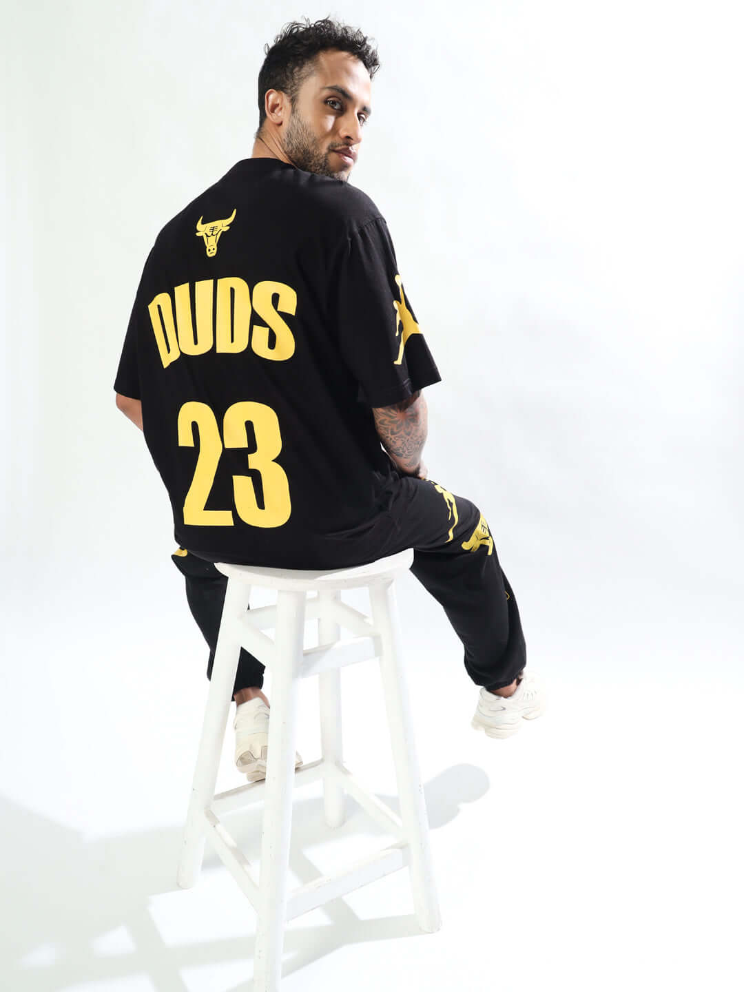 Model wearing TAURUS over-sized black t-shirt with yellow DUDS branding and the number 23, sitting on a white stool.