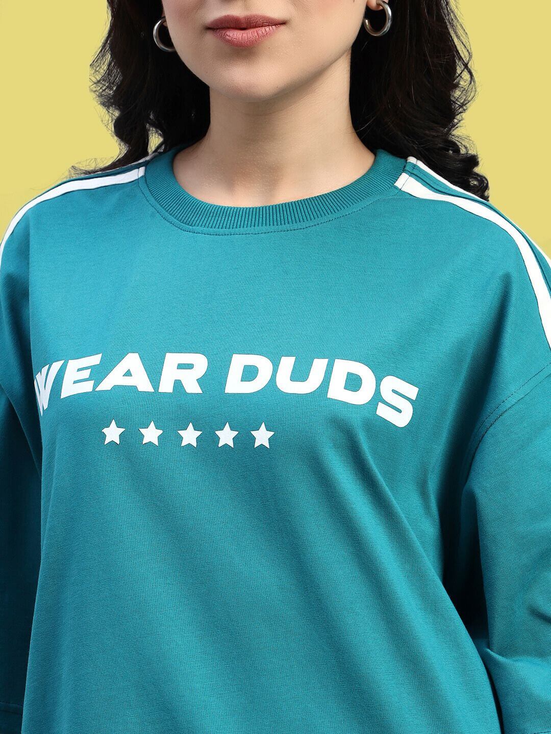 Close-up of WOMEN'S STAR JUMPER OVER-SIZED T-SHIRT TEAL showcasing star details and bold lettering on teal fabric against a yellow background.