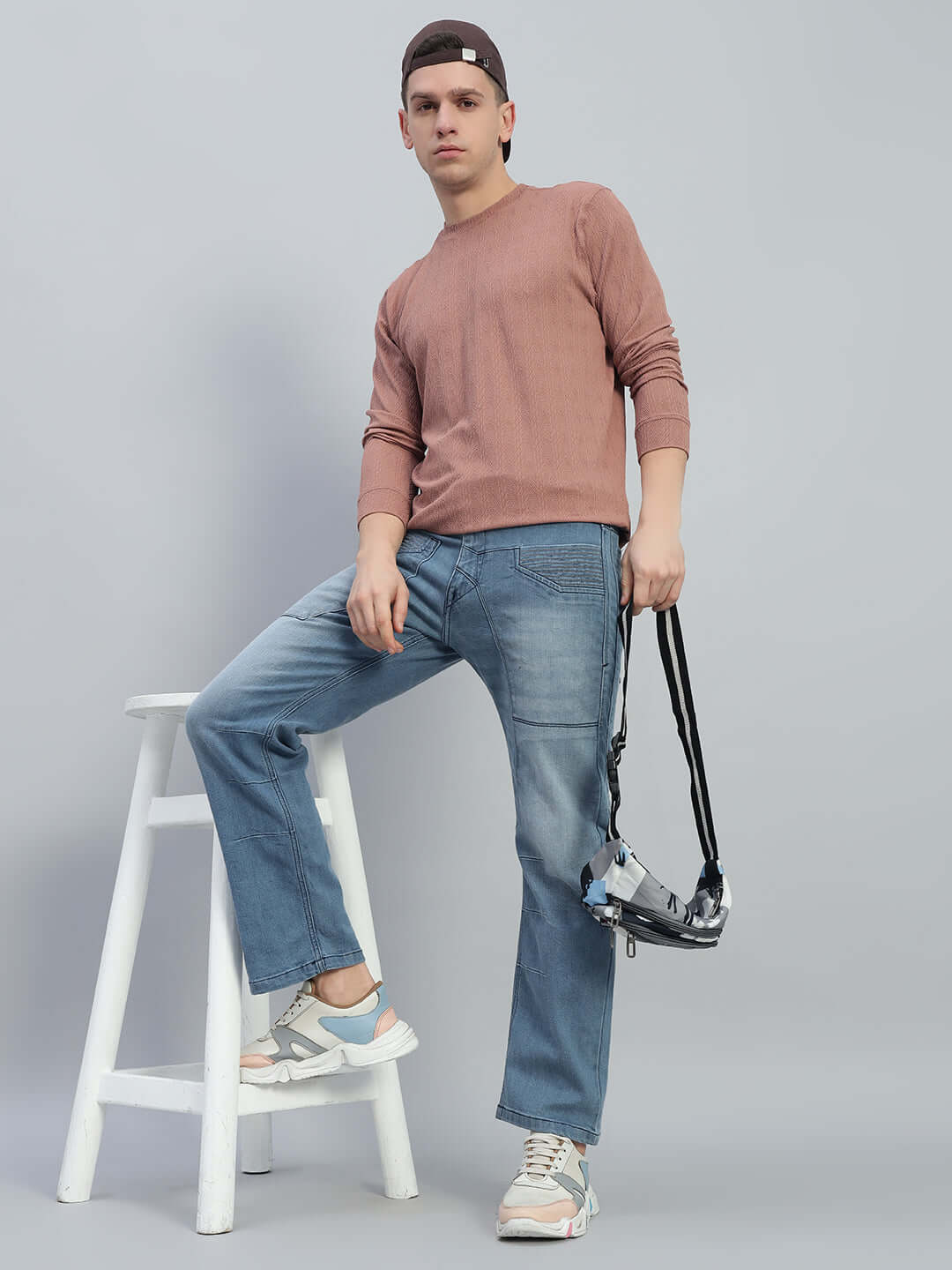 Model wearing Thrifty Regular Fit Denim Jeans in blue, paired with a casual pink sweater and sneakers, leaning on a white stool.