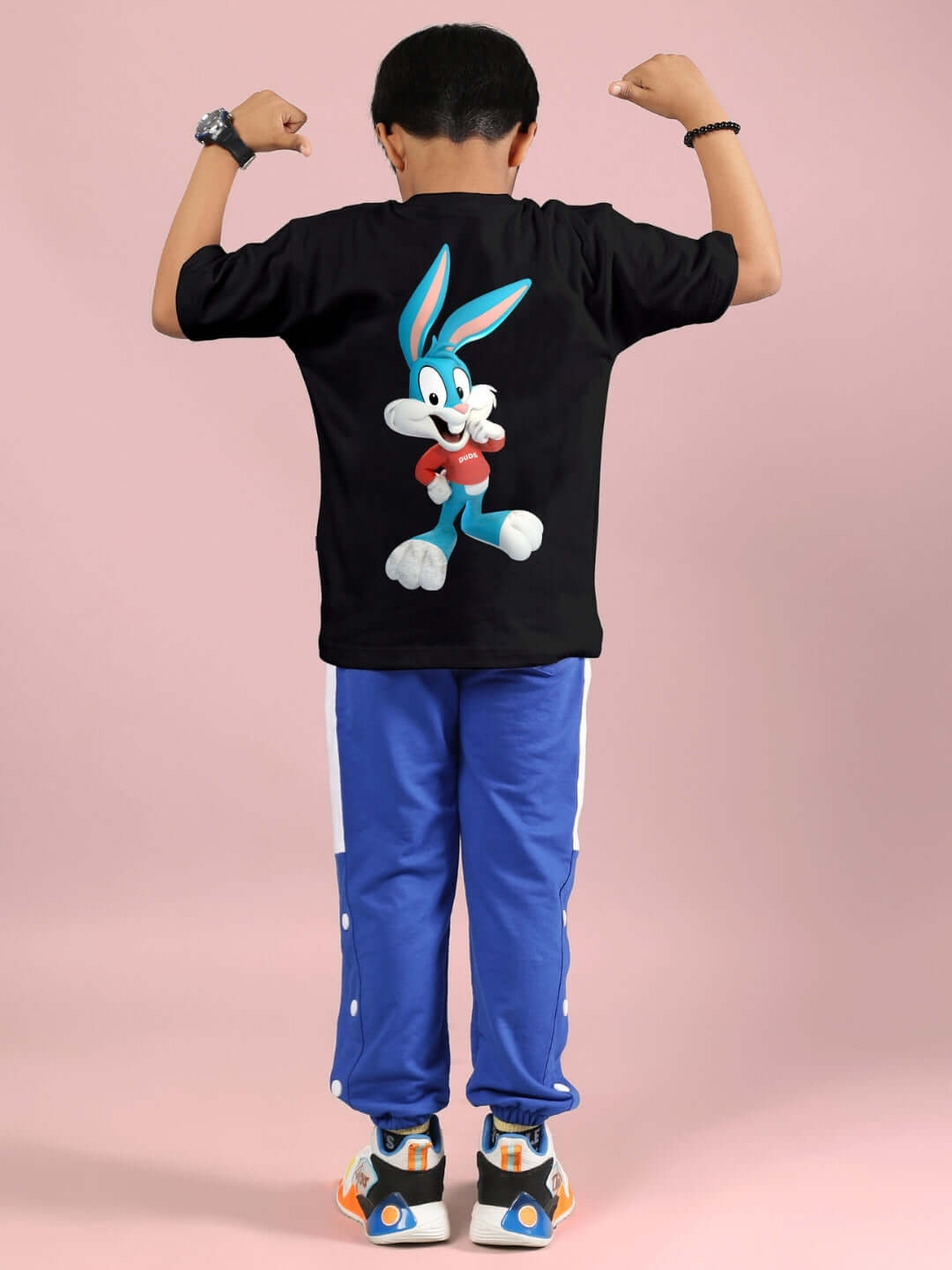 Child wearing Tiny Toon printed black and blue co-ord set, featuring a character on the back of the shirt, posing on a pink background.