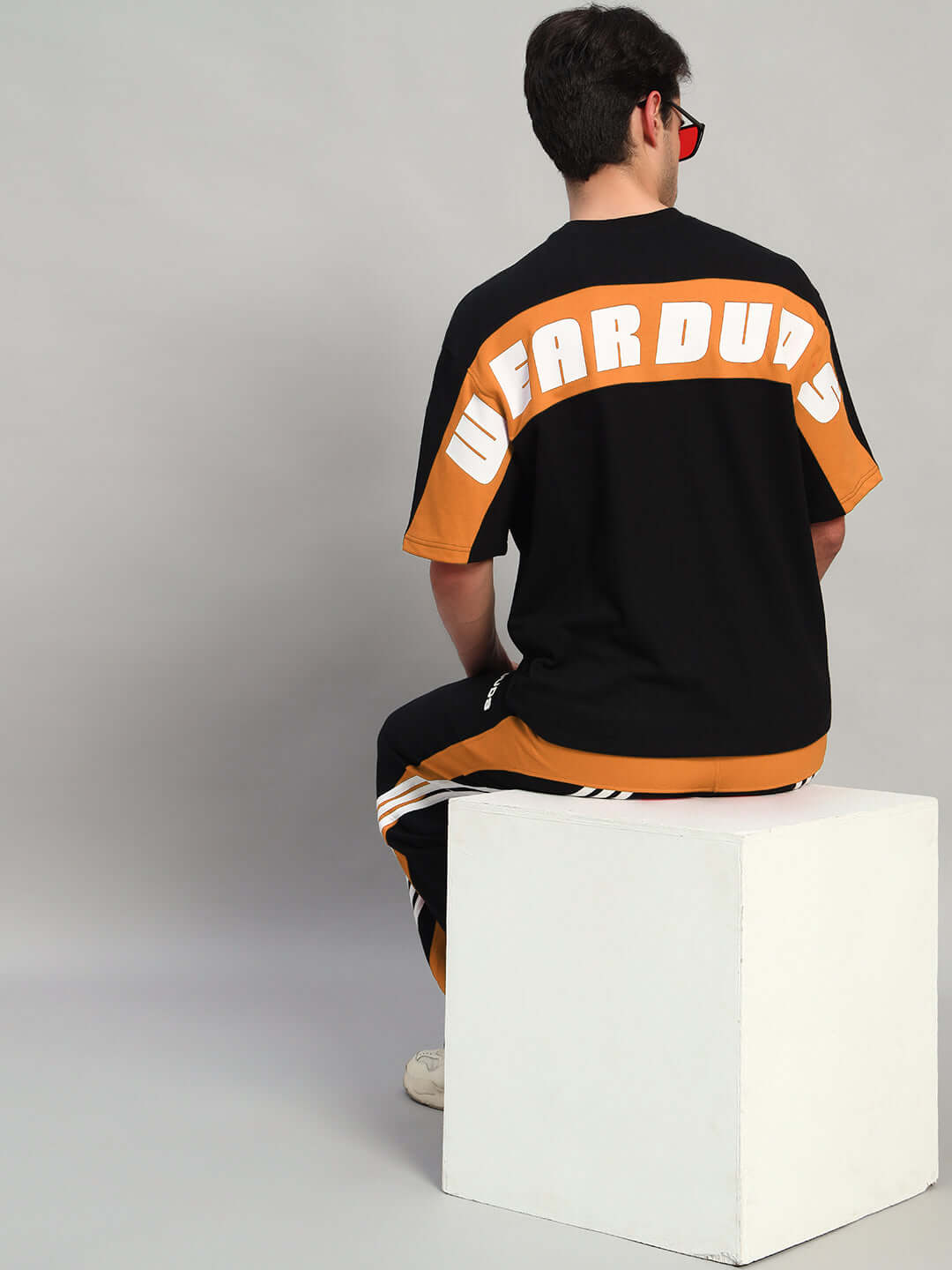 Person wearing TITAN OVER-SIZED T-SHIRT BLACK-ORANGE sitting on a white cube, showcasing the back design in a studio setting.