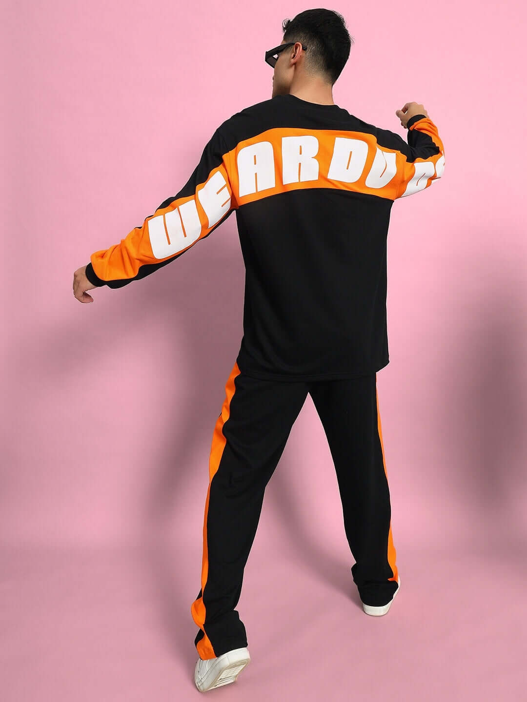 Person wearing black and orange 'WEARDO' co-ord set with white sneakers, posing against a pink background.