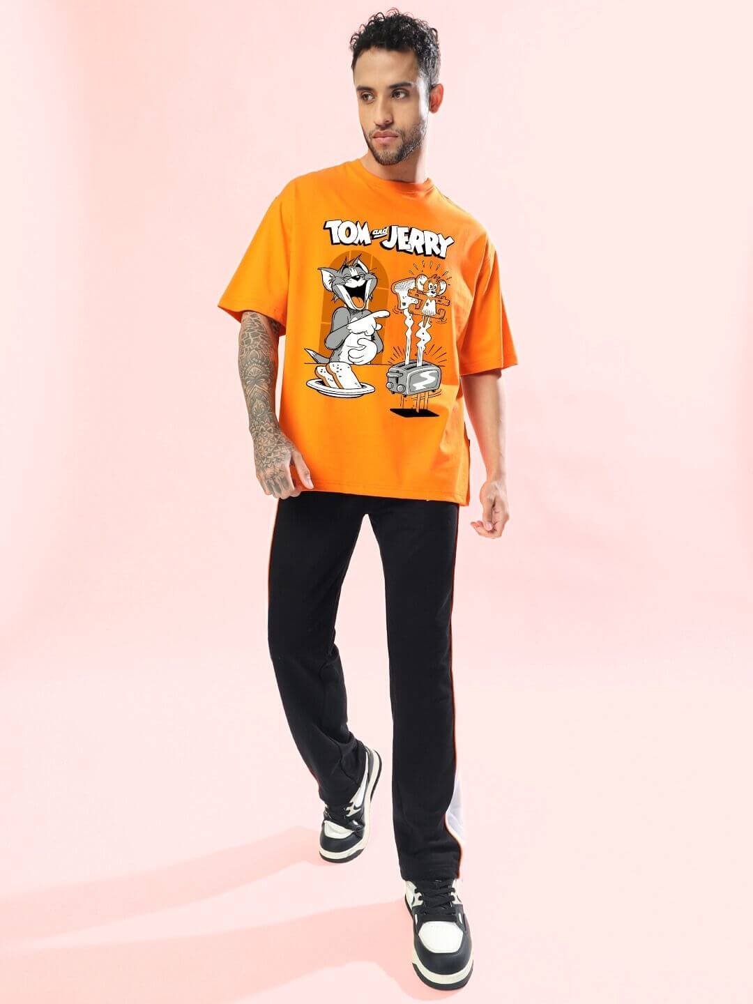Man wearing TOM AND JERRY TOAST OVERSIZED T-SHIRT ORANGE with playful cartoon graphic, standing stylishly against a pink background.