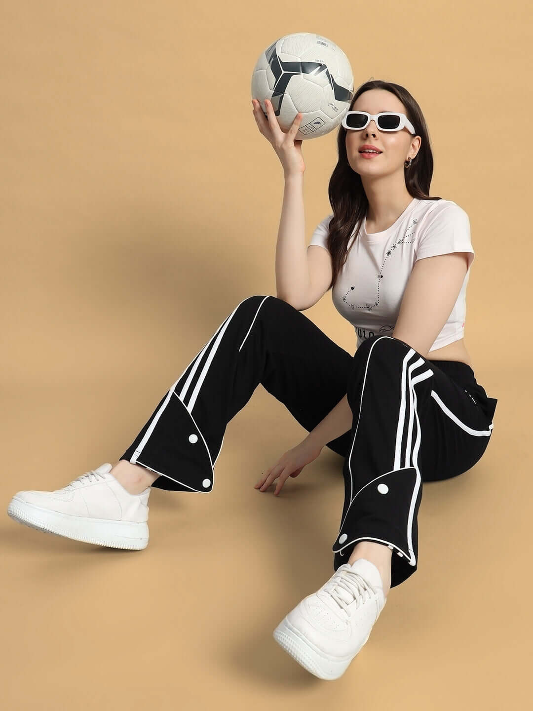 Woman in stylish outfit with sunglasses sitting on floor holding soccer ball, wearing white sneakers and black pants with white stripes.
