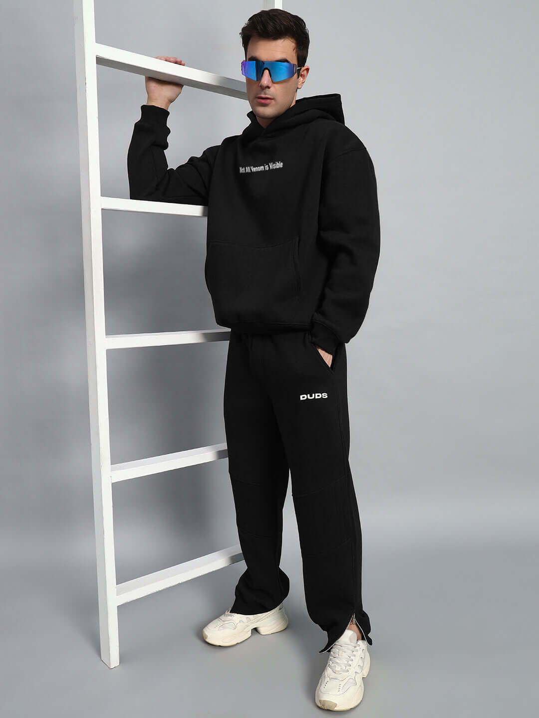 Model wearing a TRUSTISSUE oversized black hoodie, styled with a matching DUDS black sweatpants and white sneakers, against a minimalist backdrop.