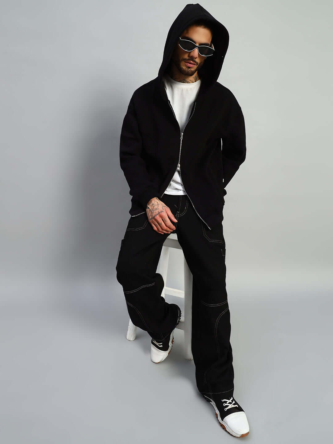 Model wearing WEARDUDS TWO FOLD ZIPPER HOODIE BLACK, sitting on stool, showcasing the hoodie design in a casual setting.